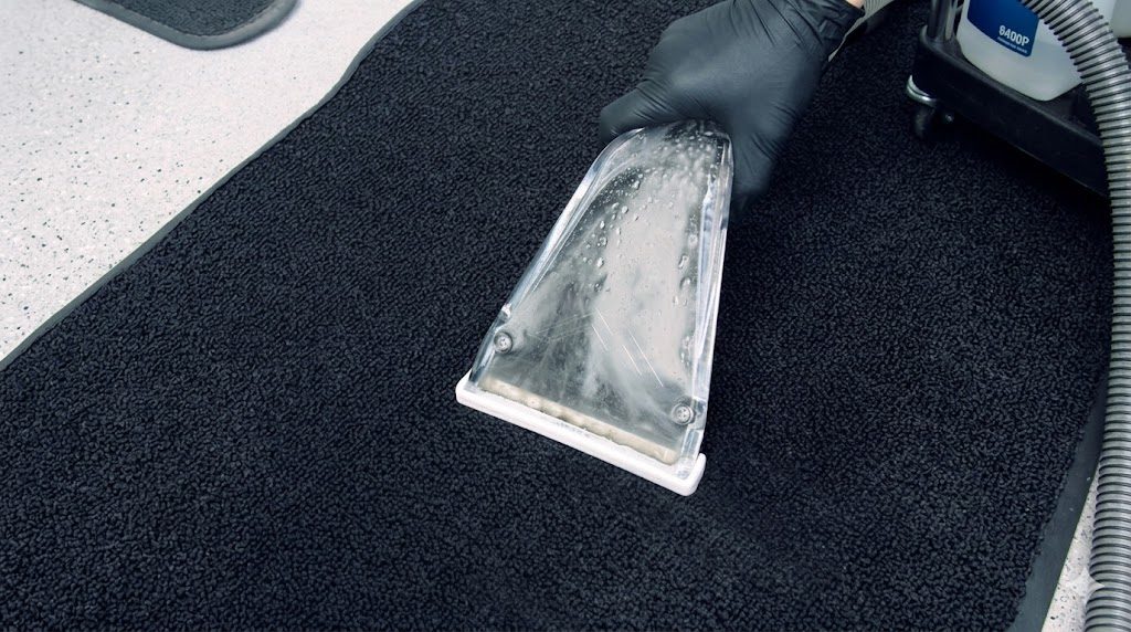 Gloved After Hours detailer drawing a hot-water extraction wand across a black car floor mat, water lifting up through the clear wand head — deep extraction that pulls ground-in dirt and salt out of the carpet fibers, not just the surface.